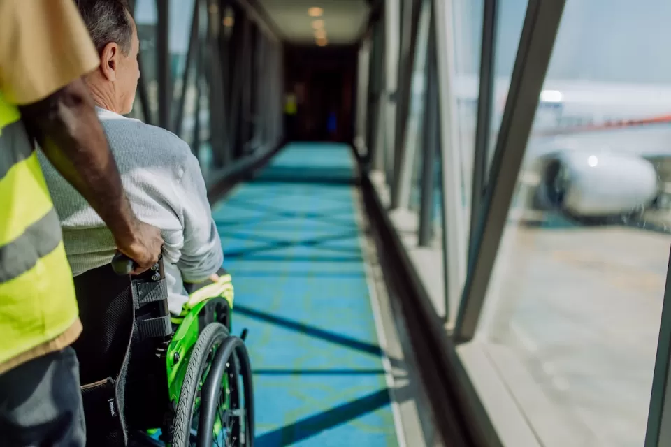 Person pusing person sitting in wheelchair down airplane ramp toward airplane at airport.